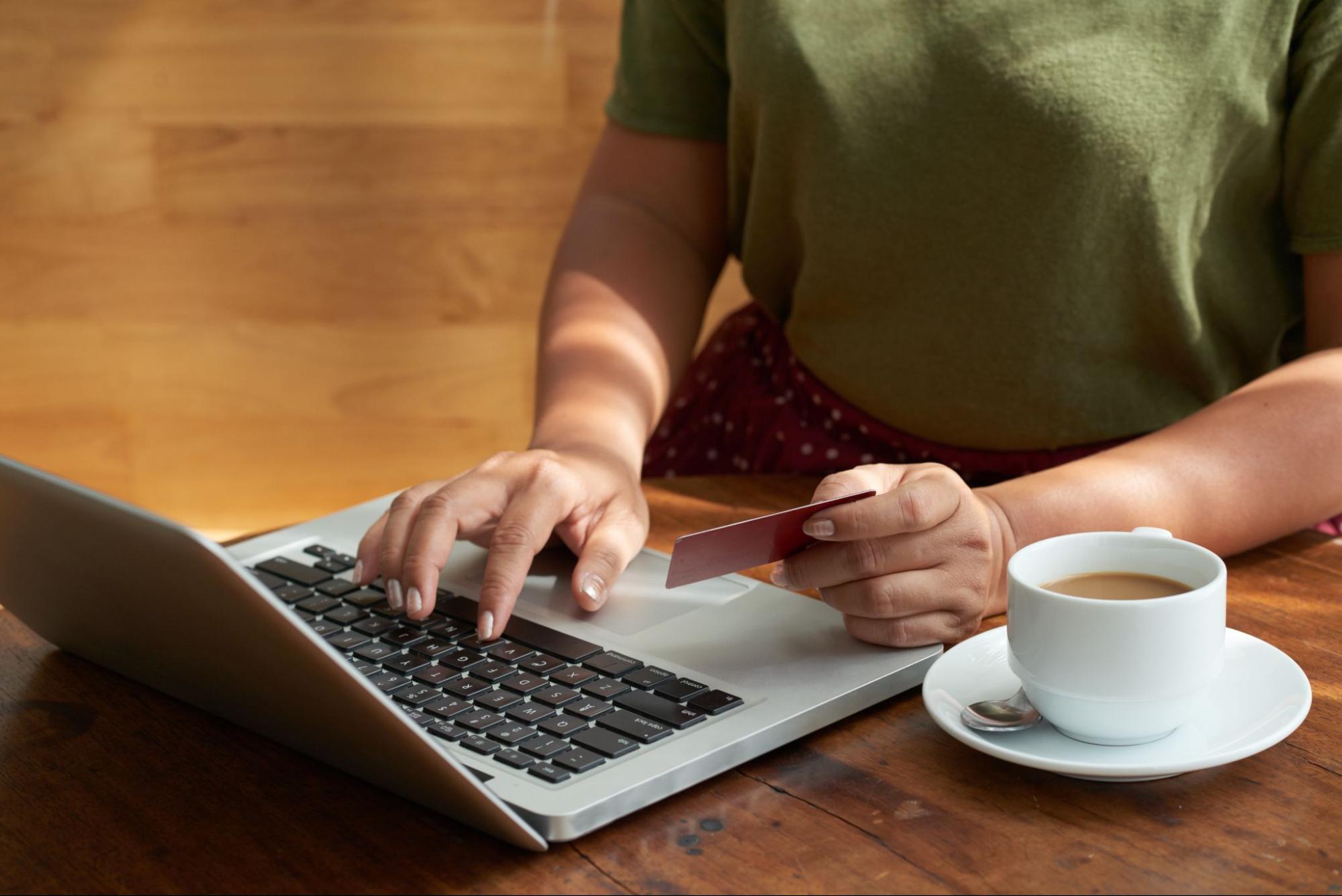 Personne utilisant un ordinateur portable avec une carte bancaire à la main et une tasse de café à proximité.